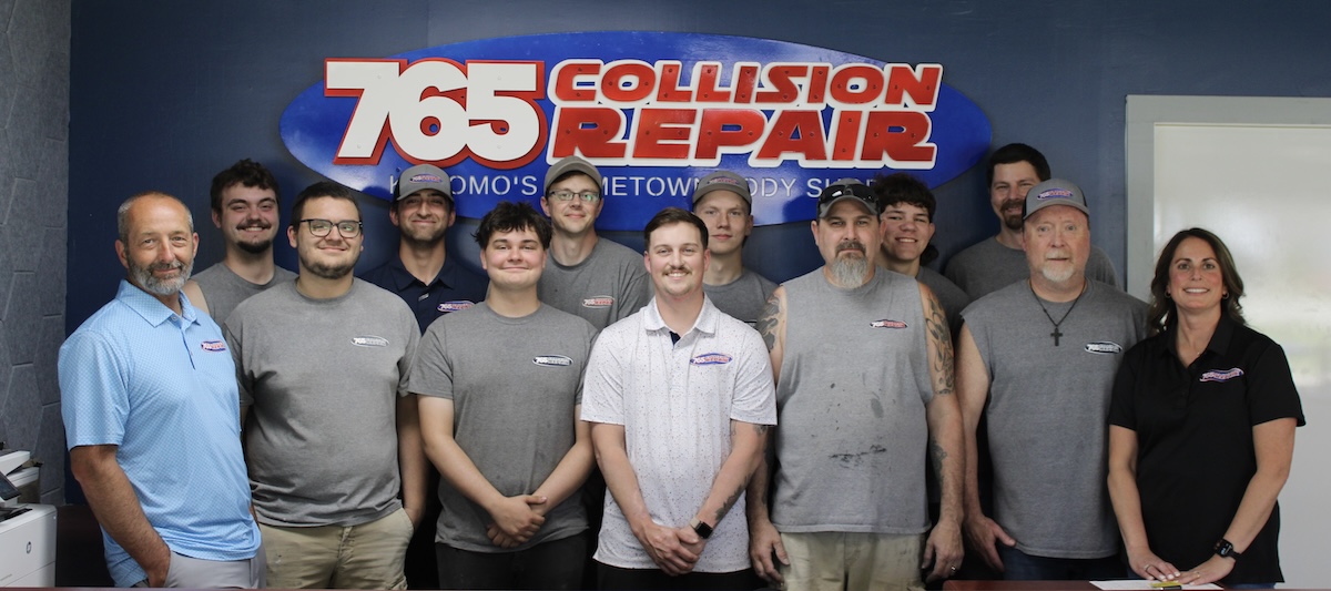 A group photo of twelve employees standing in front of a blue wall inside an office. A large, prominent oval sign behind them reads "765 Collision Repair" in red and white lettering. The team consists of eleven men and one woman, mostly wearing grey sleeveless shirts or t-shirts with a small logo, while a few wear polo shirts or hats. They are lined up side-by-side, smiling directly at the camera.