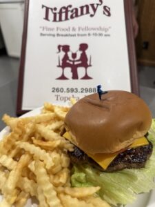 A close-up shot of a cheeseburger and a side of crinkle-cut french fries on a white plate. The cheeseburger features a grilled beef patty, melted cheddar cheese, a tomato slice, lettuce, and a brioche bun held together with a blue plastic toothpick. In the background, a white tabletop sign displays the Tiffany's restaurant logo, the slogan "Fine Food & Fellowship", the phone number 260.593.2988, and "Topeka, IN".h