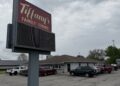 A tall, gray roadside marquee sign with a maroon top section reading "Tiffany's FAMILY DINING" sits in front of a busy restaurant parking lot. The restaurant is a single-story building with gray siding and a brown roof. Several parked cars, SUVs, and pickup trucks fill the asphalt lot under a cloudy, overcast gray sky.