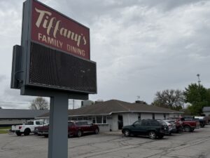 A tall, gray roadside marquee sign with a maroon top section reading "Tiffany's FAMILY DINING" sits in front of a busy restaurant parking lot. The restaurant is a single-story building with gray siding and a brown roof. Several parked cars, SUVs, and pickup trucks fill the asphalt lot under a cloudy, overcast gray sky.