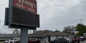 A tall, gray roadside marquee sign with a maroon top section reading "Tiffany's FAMILY DINING" sits in front of a busy restaurant parking lot. The restaurant is a single-story building with gray siding and a brown roof. Several parked cars, SUVs, and pickup trucks fill the asphalt lot under a cloudy, overcast gray sky.