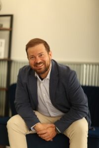 A portrait of a smiling man with a beard and brown hair, wearing a navy blue blazer, a light-colored button-up shirt, and khaki pants, leaning forward while sitting on a dark blue couch in an office setting.