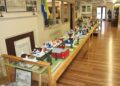A detailed miniature diorama of a historical town called Middletown is displayed on a long wooden table inside a museum. The scale model features tiny, colorful houses, barns, and trees arranged along a modeled road. The display is positioned next to a brick fireplace mantle, with informational signs, a museum display case, and a polished wood floor extending into the background.