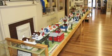 A detailed miniature diorama of a historical town called Middletown is displayed on a long wooden table inside a museum. The scale model features tiny, colorful houses, barns, and trees arranged along a modeled road. The display is positioned next to a brick fireplace mantle, with informational signs, a museum display case, and a polished wood floor extending into the background.