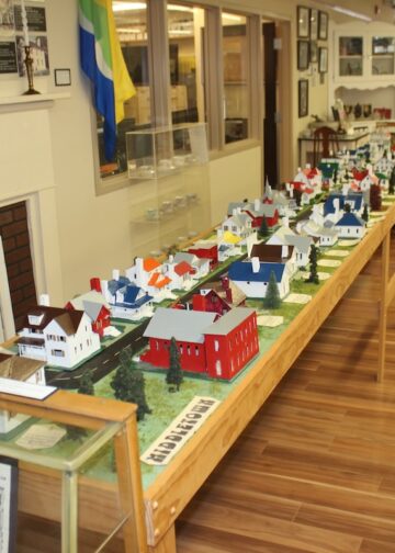 A detailed miniature diorama of a historical town called Middletown is displayed on a long wooden table inside a museum. The scale model features tiny, colorful houses, barns, and trees arranged along a modeled road. The display is positioned next to a brick fireplace mantle, with informational signs, a museum display case, and a polished wood floor extending into the background.