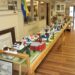 A detailed miniature diorama of a historical town called Middletown is displayed on a long wooden table inside a museum. The scale model features tiny, colorful houses, barns, and trees arranged along a modeled road. The display is positioned next to a brick fireplace mantle, with informational signs, a museum display case, and a polished wood floor extending into the background.