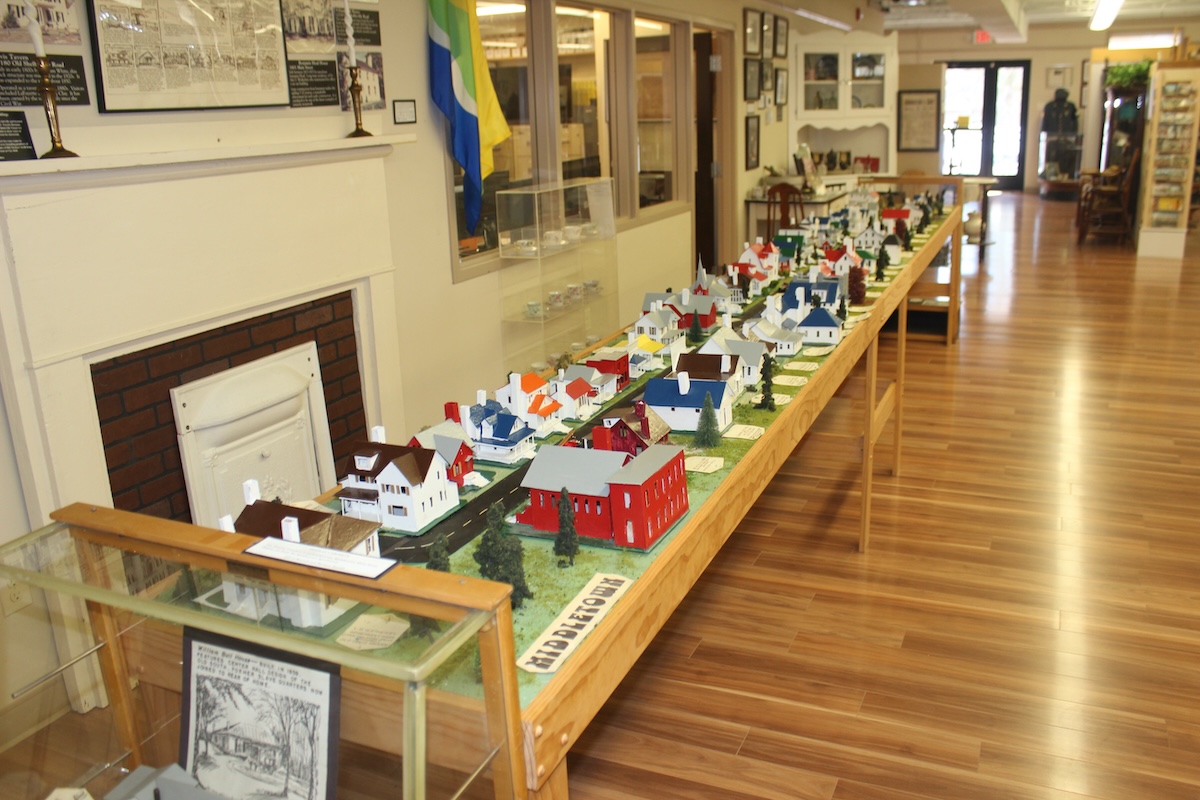 A detailed miniature diorama of a historical town called Middletown is displayed on a long wooden table inside a museum. The scale model features tiny, colorful houses, barns, and trees arranged along a modeled road. The display is positioned next to a brick fireplace mantle, with informational signs, a museum display case, and a polished wood floor extending into the background.
