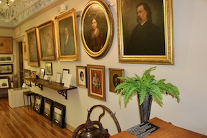 A long hallway gallery inside a museum displays a collection of historical oil painting portraits mounted on a light-colored wall. The portraits features individuals from the 19th century in ornate gold frames, arranged above a low display shelf and smaller framed artifacts. In the foreground, a vibrant green fern sits in a dark vase on a wooden surface, with a polished wood floor stretching down the corridor.