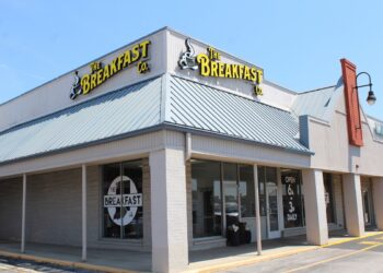 A corner storefront of "The Breakfast Co." restaurant in a strip mall under a clear blue sky. The building features light beige brick walls, large glass windows, and a prominent blue metal awning roof. Yellow neon-style signs displaying the restaurant's name and a steaming coffee cup logo are mounted on the roof line. A window decal shows the restaurant name inside an outline of the state of Indiana, and a sign near the door notes they are open from 6:00 AM to 3:00 PM daily.