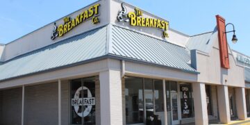A corner storefront of "The Breakfast Co." restaurant in a strip mall under a clear blue sky. The building features light beige brick walls, large glass windows, and a prominent blue metal awning roof. Yellow neon-style signs displaying the restaurant's name and a steaming coffee cup logo are mounted on the roof line. A window decal shows the restaurant name inside an outline of the state of Indiana, and a sign near the door notes they are open from 6:00 AM to 3:00 PM daily.
