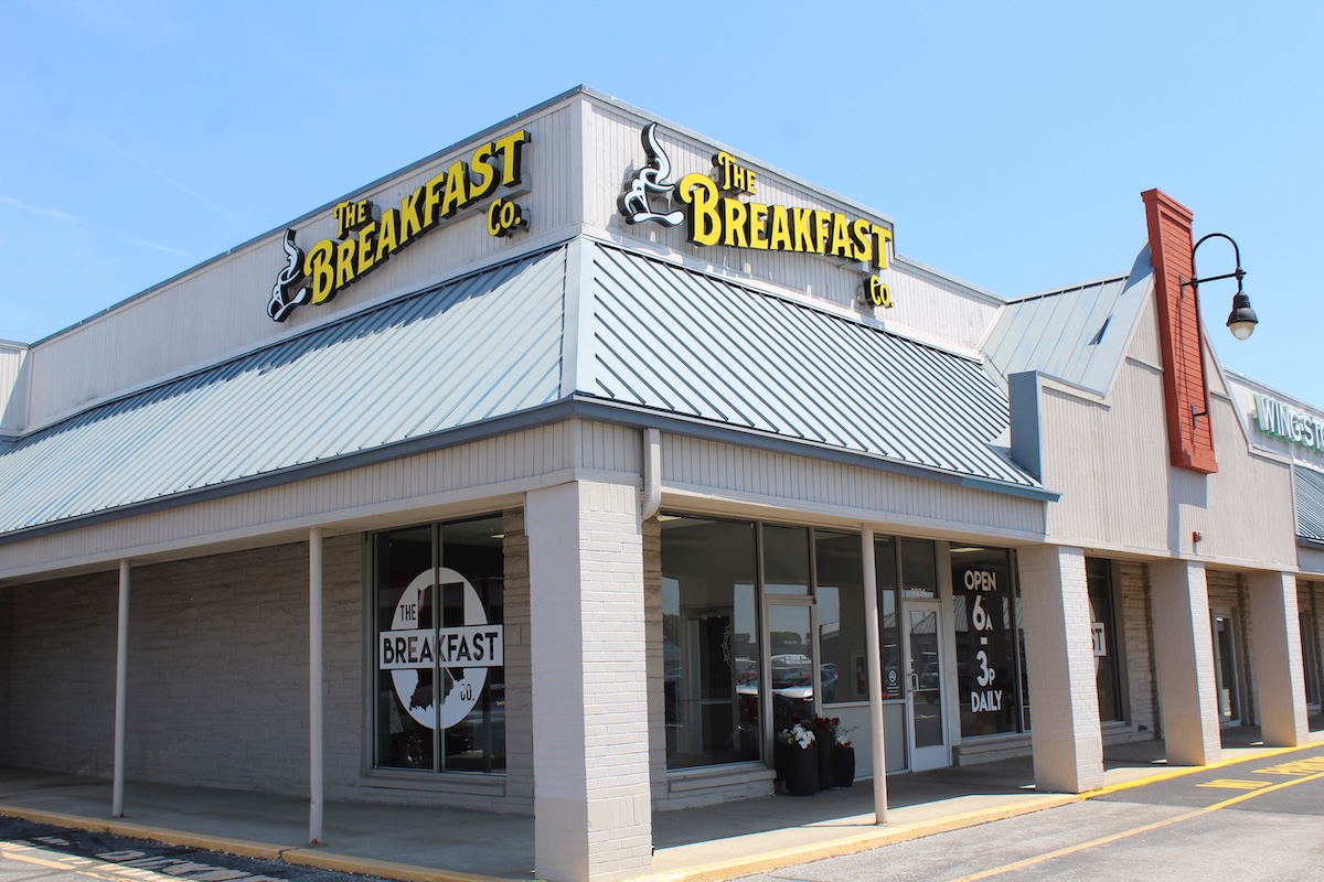 A corner storefront of "The Breakfast Co." restaurant in a strip mall under a clear blue sky. The building features light beige brick walls, large glass windows, and a prominent blue metal awning roof. Yellow neon-style signs displaying the restaurant's name and a steaming coffee cup logo are mounted on the roof line. A window decal shows the restaurant name inside an outline of the state of Indiana, and a sign near the door notes they are open from 6:00 AM to 3:00 PM daily.