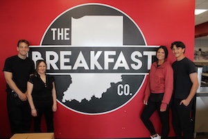 Four smiling employees pose together in front of a large red wall. The wall features a big black-and-white circular logo for "The Breakfast Co." with an outline of the state of Indiana in the center. On the left side stand a man and a woman dressed in all-black attire. On the right side stand a woman wearing a mauve hoodie with black pants and a man wearing a black t-shirt.