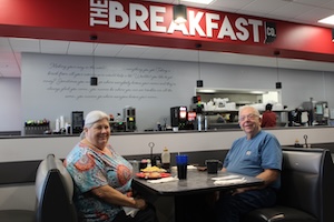 An elderly couple sits smiling in a dark booth inside The Breakfast Co. restaurant. A woman with short white hair wearing a colorful patterned shirt sits on the left. A man wearing a blue t-shirt and glasses sits on the right. Their table holds drinks and a plate of food. In the background, a large red soffit displays the restaurant's name in bold white text above a kitchen service window and a wall with cursive script.