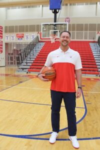 A smiling man stands alone on a polished wooden basketball court, facing the camera. He wears a short-sleeved athletic polo shirt that is white on top and red on the bottom, dark pants, and white sneakers. He holds a brown basketball under his right arm. The background shows empty red gymnasium bleachers, stairs, a basketball hoop, and bright indoor gym lighting.
