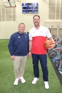 Two men stand side-by-side smiling on green indoor turf. The man on the left wears a navy blue long-sleeved Nike sweatshirt with a red cursive logo, khaki pants, and white running shoes. The man on the right wears a short-sleeved athletic polo shirt that is white on top and red on the bottom, dark pants, and white sneakers while holding a basketball under his left arm. A digital scoreboard displaying "Quakers" and "Guest" hangs on the wall behind them above a weightlifting area.
