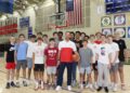 A large group of teenage male basketball players poses in a gymnasium with their coach. The coach stands front and center in a white and red athletic shirt and dark pants, holding a basketball. The players stand closely behind and around him on the polished wooden basketball court, wearing various athletic shirts, including Plainfield basketball gear. An American flag hangs on the brick wall in the background between two basketball hoops, next to sports banners and a scoreboard.