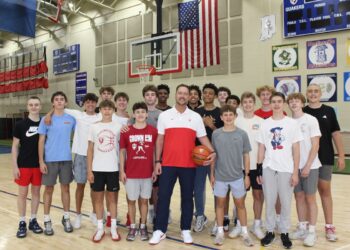 A large group of teenage male basketball players poses in a gymnasium with their coach. The coach stands front and center in a white and red athletic shirt and dark pants, holding a basketball. The players stand closely behind and around him on the polished wooden basketball court, wearing various athletic shirts, including Plainfield basketball gear. An American flag hangs on the brick wall in the background between two basketball hoops, next to sports banners and a scoreboard.