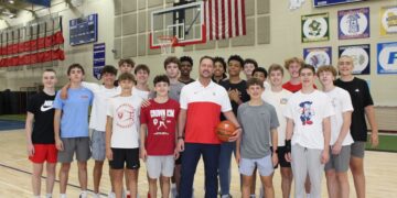 A large group of teenage male basketball players poses in a gymnasium with their coach. The coach stands front and center in a white and red athletic shirt and dark pants, holding a basketball. The players stand closely behind and around him on the polished wooden basketball court, wearing various athletic shirts, including Plainfield basketball gear. An American flag hangs on the brick wall in the background between two basketball hoops, next to sports banners and a scoreboard.