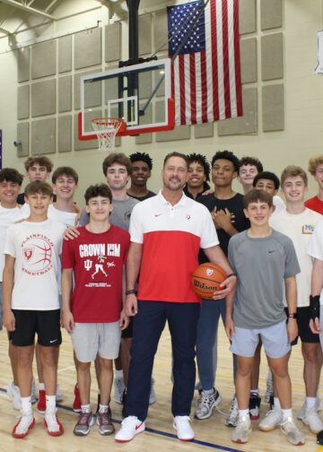 A large group of teenage male basketball players poses in a gymnasium with their coach. The coach stands front and center in a white and red athletic shirt and dark pants, holding a basketball. The players stand closely behind and around him on the polished wooden basketball court, wearing various athletic shirts, including Plainfield basketball gear. An American flag hangs on the brick wall in the background between two basketball hoops, next to sports banners and a scoreboard.