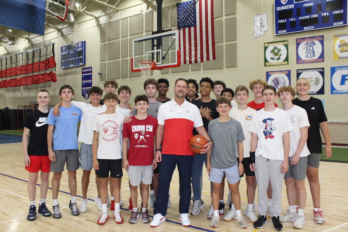A large group of teenage male basketball players poses in a gymnasium with their coach. The coach stands front and center in a white and red athletic shirt and dark pants, holding a basketball. The players stand closely behind and around him on the polished wooden basketball court, wearing various athletic shirts, including Plainfield basketball gear. An American flag hangs on the brick wall in the background between two basketball hoops, next to sports banners and a scoreboard.