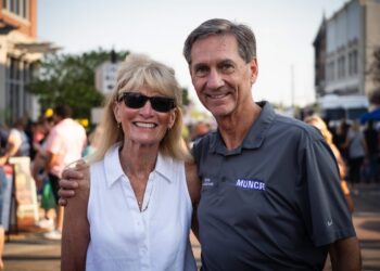 A smiling, middle-aged man and woman pose closely together for a portrait on a crowded outdoor city street during the day. The man on the right, Muncie Mayor Dan Ridenour, has brown hair and wears a gray Nike polo shirt printed with the word "MUNCIE" on the chest. He has his arm around the woman on the left, Sherry Ridenour, who is wearing sunglasses, a sleeveless white button-down shirt, and a necklace. The blurred background shows a street event with numerous pedestrians and buildings under bright sunlight.