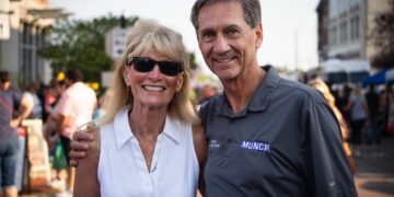 A smiling, middle-aged man and woman pose closely together for a portrait on a crowded outdoor city street during the day. The man on the right, Muncie Mayor Dan Ridenour, has brown hair and wears a gray Nike polo shirt printed with the word "MUNCIE" on the chest. He has his arm around the woman on the left, Sherry Ridenour, who is wearing sunglasses, a sleeveless white button-down shirt, and a necklace. The blurred background shows a street event with numerous pedestrians and buildings under bright sunlight.