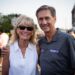 A smiling, middle-aged man and woman pose closely together for a portrait on a crowded outdoor city street during the day. The man on the right, Muncie Mayor Dan Ridenour, has brown hair and wears a gray Nike polo shirt printed with the word "MUNCIE" on the chest. He has his arm around the woman on the left, Sherry Ridenour, who is wearing sunglasses, a sleeveless white button-down shirt, and a necklace. The blurred background shows a street event with numerous pedestrians and buildings under bright sunlight.