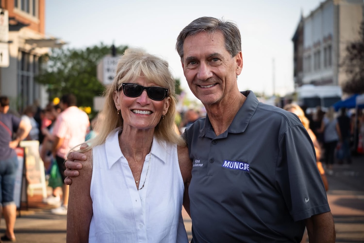 A smiling, middle-aged man and woman pose closely together for a portrait on a crowded outdoor city street during the day. The man on the right, Muncie Mayor Dan Ridenour, has brown hair and wears a gray Nike polo shirt printed with the word "MUNCIE" on the chest. He has his arm around the woman on the left, Sherry Ridenour, who is wearing sunglasses, a sleeveless white button-down shirt, and a necklace. The blurred background shows a street event with numerous pedestrians and buildings under bright sunlight.