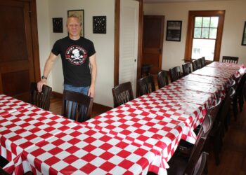A man wearing a black "Greek’s Pizzeria" graphic t-shirt stands proudly at the head of a large, U-shaped banquet table arrangement inside a warm, wood-accented dining room. The long wooden tables are neatly dressed in classic red-and-white checkered tablecloths and lined with dark wooden chairs. Polished hardwood floors, historic dark wood trim, windows, and framed art pieces decorate the surrounding walls, creating a welcoming private event space.