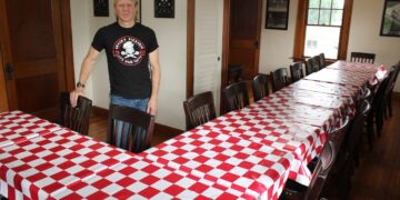 A man wearing a black "Greek’s Pizzeria" graphic t-shirt stands proudly at the head of a large, U-shaped banquet table arrangement inside a warm, wood-accented dining room. The long wooden tables are neatly dressed in classic red-and-white checkered tablecloths and lined with dark wooden chairs. Polished hardwood floors, historic dark wood trim, windows, and framed art pieces decorate the surrounding walls, creating a welcoming private event space.