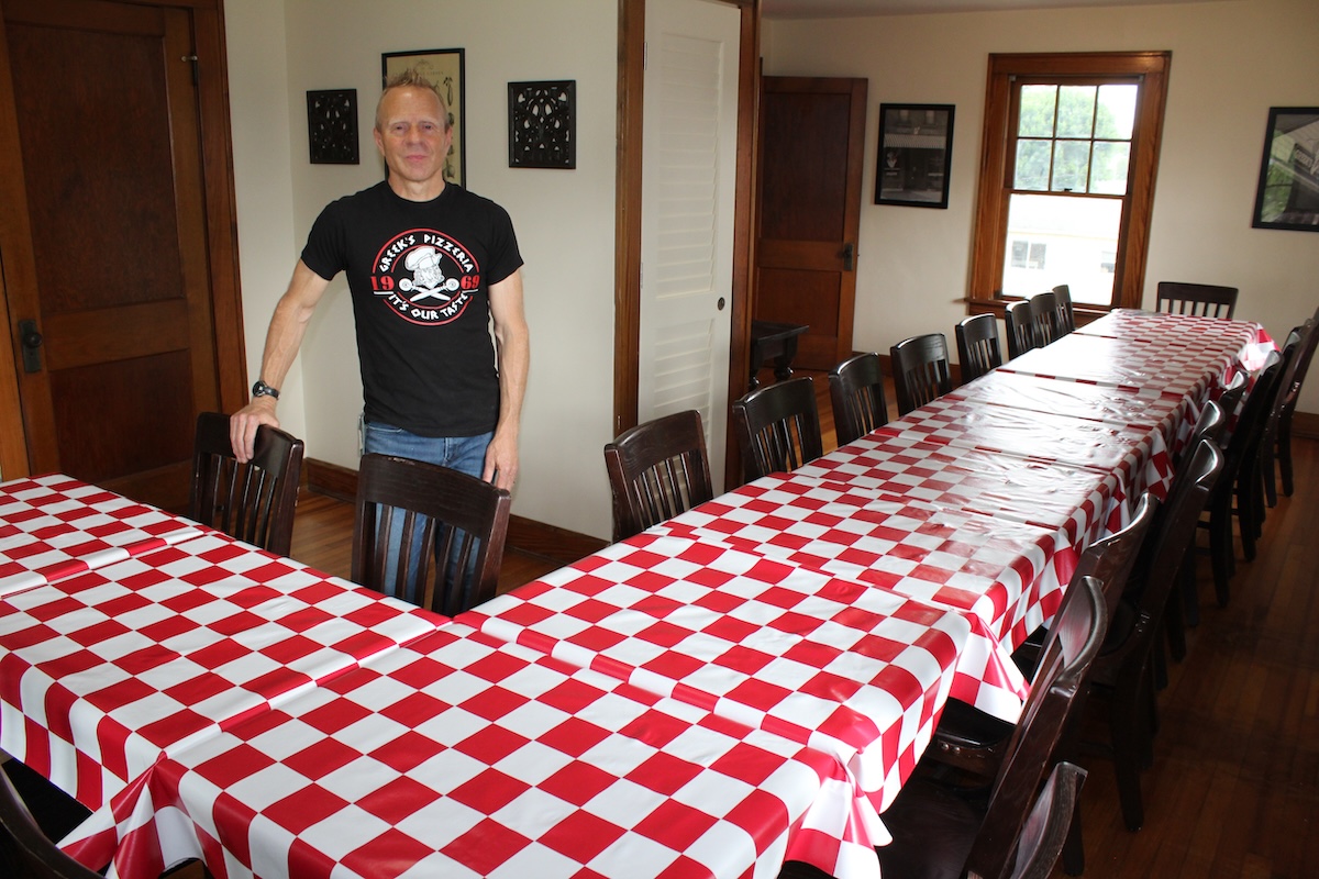 A man wearing a black "Greek’s Pizzeria" graphic t-shirt stands proudly at the head of a large, U-shaped banquet table arrangement inside a warm, wood-accented dining room. The long wooden tables are neatly dressed in classic red-and-white checkered tablecloths and lined with dark wooden chairs. Polished hardwood floors, historic dark wood trim, windows, and framed art pieces decorate the surrounding walls, creating a welcoming private event space.