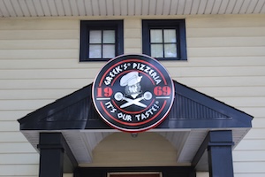 A symmetrical exterior view displays a large, circular "Greek’s Pizzeria" sign mounted onto the dark gable above the building's main entrance portico. The sign features the cartoon mascot chef, the founding year "1969," and the slogan "IT'S OUR TASTE!" on a black background bordered in red and white. Two small square windows with dark frames sit directly above the sign on the cream-colored horizontal lap siding of the house structure.