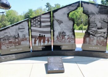 A black granite monument consists of four large vertical panels split into two sections, with a central cutout silhouette of a saluting soldier. The panels are etched with detailed black-and-white scenes and titles at the bottom: "HOMELAND" shows a community parade; "FAMILY" displays silhouettes of a family holding hands before an American flag; "PATRIOT" depicts the iconic Iwo Jima flag-raising; and "SACRIFICE" portrays a military cemetery and a grieving figure. A descriptive plaque sits on the concrete walkway in front of the base.
