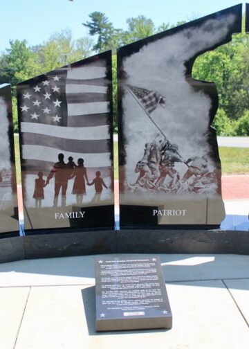 A black granite monument consists of four large vertical panels split into two sections, with a central cutout silhouette of a saluting soldier. The panels are etched with detailed black-and-white scenes and titles at the bottom: "HOMELAND" shows a community parade; "FAMILY" displays silhouettes of a family holding hands before an American flag; "PATRIOT" depicts the iconic Iwo Jima flag-raising; and "SACRIFICE" portrays a military cemetery and a grieving figure. A descriptive plaque sits on the concrete walkway in front of the base.