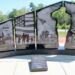 A black granite monument consists of four large vertical panels split into two sections, with a central cutout silhouette of a saluting soldier. The panels are etched with detailed black-and-white scenes and titles at the bottom: "HOMELAND" shows a community parade; "FAMILY" displays silhouettes of a family holding hands before an American flag; "PATRIOT" depicts the iconic Iwo Jima flag-raising; and "SACRIFICE" portrays a military cemetery and a grieving figure. A descriptive plaque sits on the concrete walkway in front of the base.