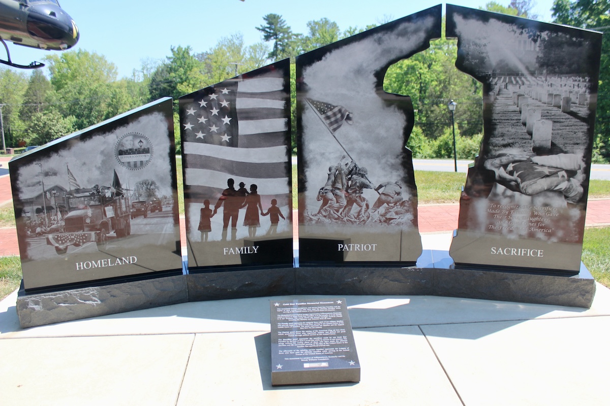 A black granite monument consists of four large vertical panels split into two sections, with a central cutout silhouette of a saluting soldier. The panels are etched with detailed black-and-white scenes and titles at the bottom: "HOMELAND" shows a community parade; "FAMILY" displays silhouettes of a family holding hands before an American flag; "PATRIOT" depicts the iconic Iwo Jima flag-raising; and "SACRIFICE" portrays a military cemetery and a grieving figure. A descriptive plaque sits on the concrete walkway in front of the base.