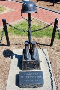 A dark bronze Battle Cross sculpture stands on a concrete base, encircled by black metal posts and chains. The traditional military memorial arrangement features a service rifle thrust downward into a pair of combat boots, with a combat helmet resting on top of the rifle stock. A small, dark stone block in front is engraved with the text "IN MEMORY OF OUR FALLEN WARRIORS, 11 NOVEMBER 2012."