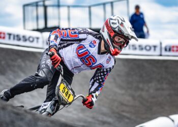 A close-up shot captures a professional BMX racing cyclist wearing a white, black, and red Team USA jersey adorned with stars and stripes leaning heavily into a turn on a paved berm. The rider wears a white and red full-face helmet with matching Oakley goggles, red racing gloves, and black pants while navigating a gray track curve. The bicycle features a bright yellow front number plate reading "541". White safety barriers with black "TISSOT" branding line the track in the soft-focus background under a cloudy blue sky.