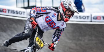 A close-up shot captures a professional BMX racing cyclist wearing a white, black, and red Team USA jersey adorned with stars and stripes leaning heavily into a turn on a paved berm. The rider wears a white and red full-face helmet with matching Oakley goggles, red racing gloves, and black pants while navigating a gray track curve. The bicycle features a bright yellow front number plate reading "541". White safety barriers with black "TISSOT" branding line the track in the soft-focus background under a cloudy blue sky.