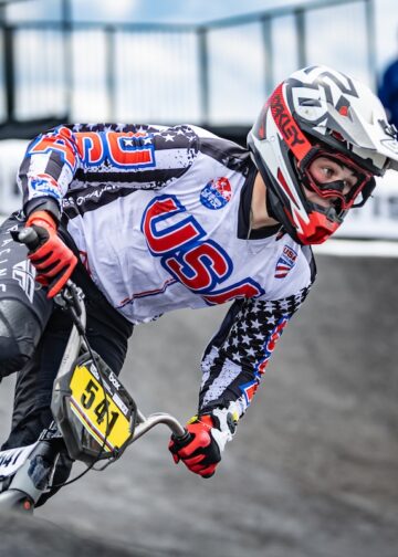 A close-up shot captures a professional BMX racing cyclist wearing a white, black, and red Team USA jersey adorned with stars and stripes leaning heavily into a turn on a paved berm. The rider wears a white and red full-face helmet with matching Oakley goggles, red racing gloves, and black pants while navigating a gray track curve. The bicycle features a bright yellow front number plate reading "541". White safety barriers with black "TISSOT" branding line the track in the soft-focus background under a cloudy blue sky.