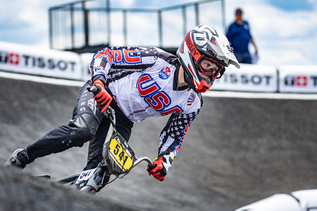 A close-up shot captures a professional BMX racing cyclist wearing a white, black, and red Team USA jersey adorned with stars and stripes leaning heavily into a turn on a paved berm. The rider wears a white and red full-face helmet with matching Oakley goggles, red racing gloves, and black pants while navigating a gray track curve. The bicycle features a bright yellow front number plate reading "541". White safety barriers with black "TISSOT" branding line the track in the soft-focus background under a cloudy blue sky.