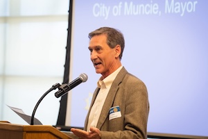 A side profile shot of Muncie Mayor Dan Ridenour speaking dynamically into a microphone at a wooden podium during a formal presentation. He wears a tan blazer over a white dress shirt and a name tag pinned to his lapel. Behind him, a large projector screen displays the text "City of Muncie, Mayor" against a light blue background.
