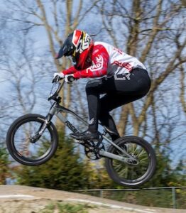 A BMX racer wearing a red, white, and black full-face helmet with Fly Racing gear is captured mid-air, catching air off a dirt track hill. The athlete leans forward over a sleek grey racing bicycle while wearing black pants and white gloves. Bare trees and a clear blue sky fill the soft-focus background behind the dirt track.