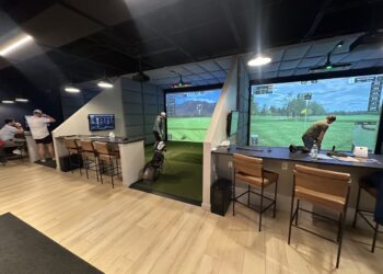 An indoor golf simulator facility with modern amenities and a clean aesthetic. The room features multiple private playing bays separated by padded gray accent walls, light wood flooring, and black acoustic ceiling tiles. In the center bay, a golfer stands on a green turf hitting mat facing a large projection screen displaying a digital golf course landscape with a mountain in the background. To the right, a second bay shows a large screen displaying a virtual golf green with data overlays. Each bay is equipped with a dark-topped bar counter, leather barstools with black metal frames, and secondary wall-mounted display monitors. In the far left background, two people stand near an additional counter area.