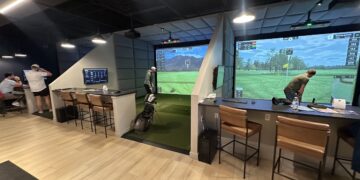 An indoor golf simulator facility with modern amenities and a clean aesthetic. The room features multiple private playing bays separated by padded gray accent walls, light wood flooring, and black acoustic ceiling tiles. In the center bay, a golfer stands on a green turf hitting mat facing a large projection screen displaying a digital golf course landscape with a mountain in the background. To the right, a second bay shows a large screen displaying a virtual golf green with data overlays. Each bay is equipped with a dark-topped bar counter, leather barstools with black metal frames, and secondary wall-mounted display monitors. In the far left background, two people stand near an additional counter area.