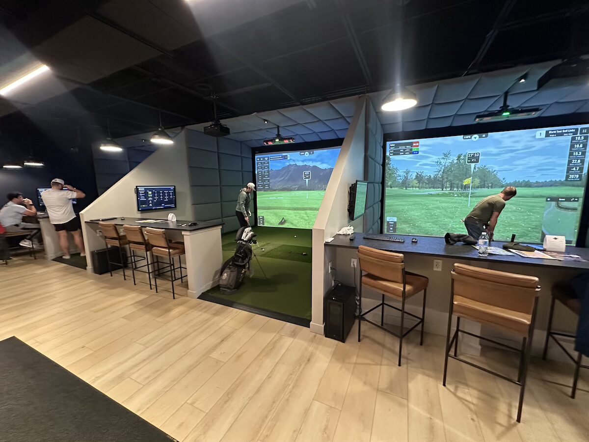 An indoor golf simulator facility with modern amenities and a clean aesthetic. The room features multiple private playing bays separated by padded gray accent walls, light wood flooring, and black acoustic ceiling tiles. In the center bay, a golfer stands on a green turf hitting mat facing a large projection screen displaying a digital golf course landscape with a mountain in the background. To the right, a second bay shows a large screen displaying a virtual golf green with data overlays. Each bay is equipped with a dark-topped bar counter, leather barstools with black metal frames, and secondary wall-mounted display monitors. In the far left background, two people stand near an additional counter area.