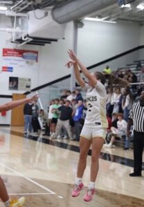A female basketball player in a white and gold uniform with "BRONCOS" and the number 2 on the jersey is captured mid-air executing a jump shot during a game. She wears pink basketball shoes and her long brown ponytail flies back behind her as she releases the ball with high extension. A referee stands on the right side of the frame, a defender's arm stretches in from the bottom left, and a crowded student section sits in the background bleachers of the indoor gymnasium.
