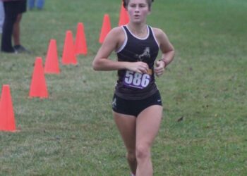 A young female athlete runs forward along a grassy cross-country course during a race. She is wearing a dark running singlet with a white trim, black athletic shorts, a competitive race bib numbered "586" on her chest, and her hair is styled in two braided buns. A line of bright orange cones marks the boundary of the course on the left. In the blurred background, spectators stand on the grass while another runner trails further behind on a cloudy day.