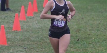 A young female athlete runs forward along a grassy cross-country course during a race. She is wearing a dark running singlet with a white trim, black athletic shorts, a competitive race bib numbered "586" on her chest, and her hair is styled in two braided buns. A line of bright orange cones marks the boundary of the course on the left. In the blurred background, spectators stand on the grass while another runner trails further behind on a cloudy day.