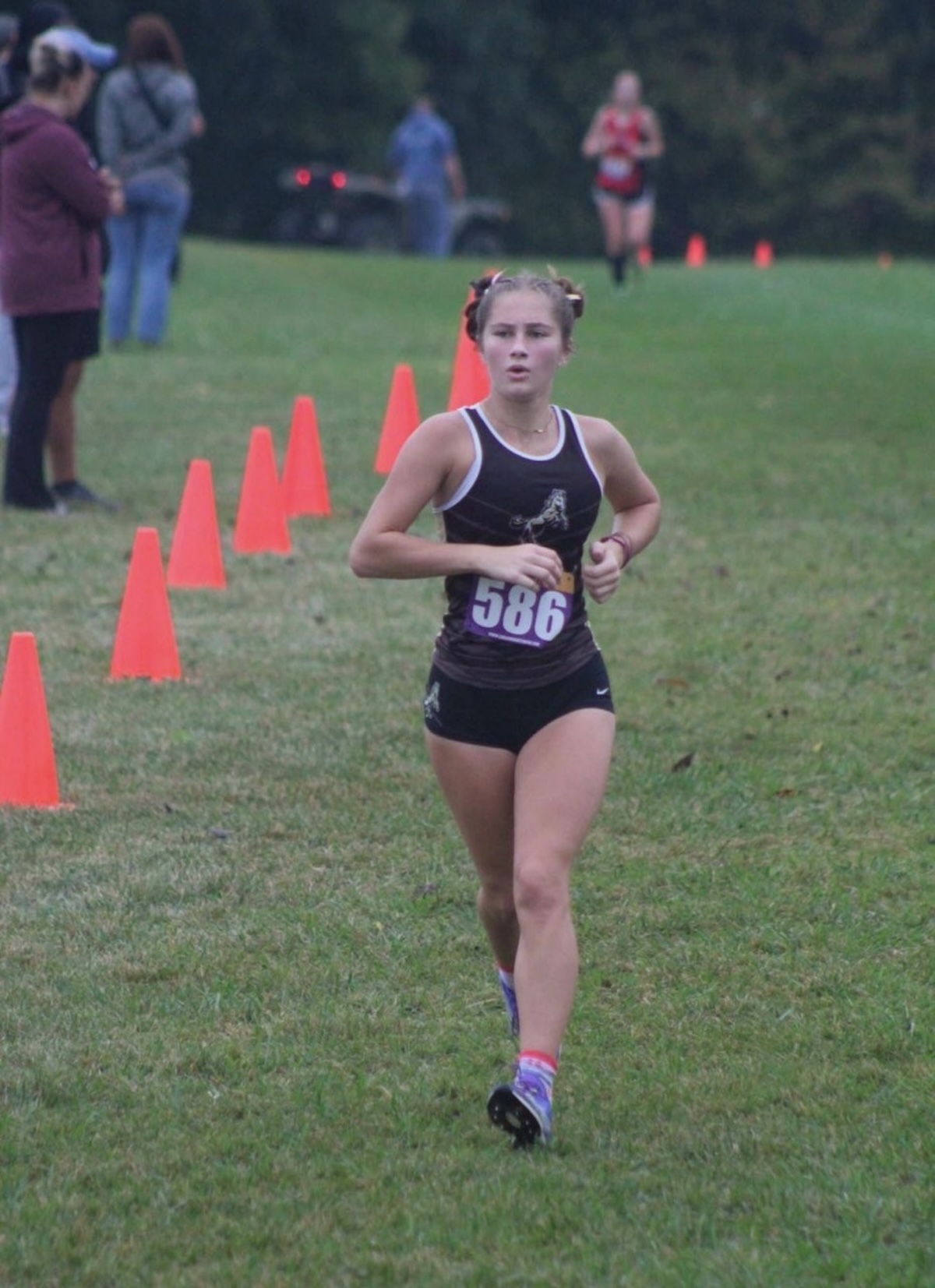 A young female athlete runs forward along a grassy cross-country course during a race. She is wearing a dark running singlet with a white trim, black athletic shorts, a competitive race bib numbered "586" on her chest, and her hair is styled in two braided buns. A line of bright orange cones marks the boundary of the course on the left. In the blurred background, spectators stand on the grass while another runner trails further behind on a cloudy day.